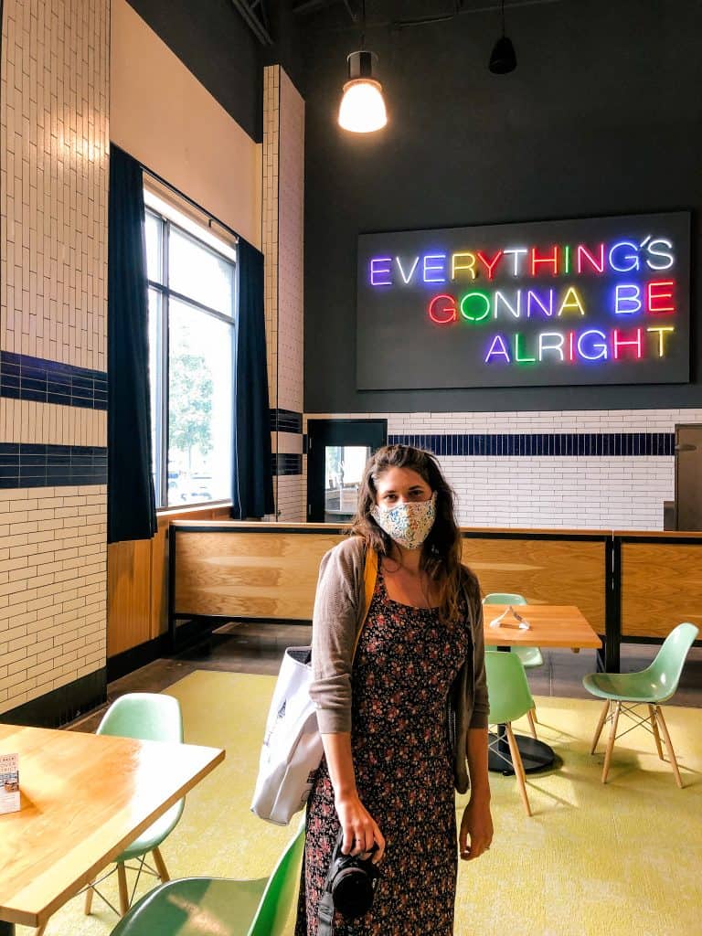 Woman in face mask in front of rainbow light up sign reading "Everything's gonna be alright"